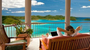 A balcony with a view of a turquoise sea and green hills, wooden furniture, and a potted plant. - Heaven's Gate - Off Grid Ocean View Villa (Coral Bay)