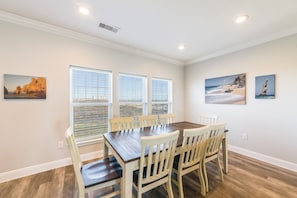 A dining room with a wooden table and white chairs, large windows with blinds, and two wall-mounted paintings.