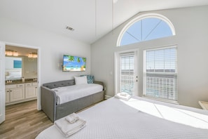 A bedroom with a large bed, a gray sofa, a TV, and a window with blinds.