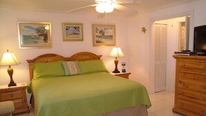 Une chambre d’hôtel comprenant un lit, deux tables de chevet avec des lampes, une commode, un ventilateur de plafond et trois tableaux encadrés accrochés au mur.