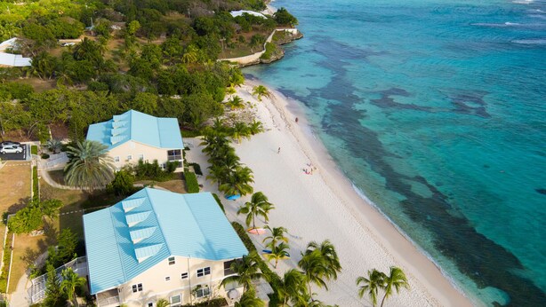Vue aérienne d’une propriété située en bord de mer, avec un grand bâtiment doté d’un toit bleu, une plage de sable et des eaux turquoise et cristallines.
