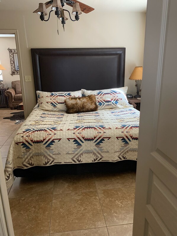 Un dormitorio con una cama, una colcha estampada, un cabecero, una lámpara y una silla al fondo.