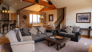 Ein geräumiges Wohnzimmer mit einem großen Ecksofa, einem Akzentstuhl mit Hounds-Tooth-Muster und einem Couchtisch. Eine Treppe führt auf eine obere Ebene, und ein Essbereich ist im Hintergrund zu sehen.