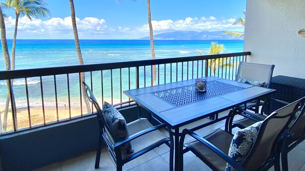 A balcony with a table and chairs overlooking a beach with palm trees and the ocean. - Direct Oceanfront Condo with Amazing Island Views!! (Lahaina)