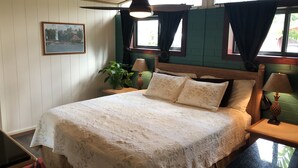 Un dormitorio con cama, dos mesitas de noche, ventilador de techo, ventana con cortinas, un cuadro colgado en la pared y una planta en maceta.