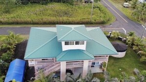 A two-story house with a green roof, a covered porch, and a small garden area with palm trees. - Nice Studio w/ A/C Near Kehena/Pohoiki Beaches and Lava Fields - sleeps 3 (Pahoa)