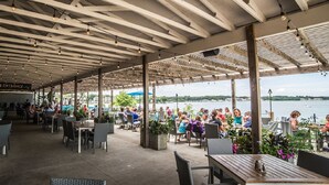 An outdoor dining area with tables and chairs, a covered ceiling with exposed beams, and a view of a body of water in the background. - Cozy Okoboji Vacation Home At Bridges Bay Resort - backs to pool! #118 (Arnolds Park)
