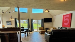 A spacious living area with a kitchen, dining area, and a large window overlooking a green landscape. - Cozy Okoboji Vacation Home At Bridges Bay Resort - backs to pool! #118 (Arnolds Park)