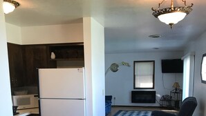 Cocina compacta con refrigerador, microondas y gabinetes de madera oscura. Se observa un comedor con una mesa de vidrio y sillas negras.
