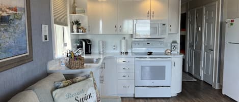 A modern kitchen with white cabinets, a microwave, and a stove. There is a basket of snacks on the counter and a decorative pillow on the couch.