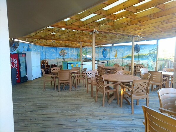 An outdoor dining area with wooden tables and chairs, a mural on the wall, and a Coca-Cola refrigerator.
