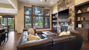 A spacious living room with a stone fireplace, leather sofa, and large windows offering a view of the outdoors. - Central Location w/ Suncadia Pool Access (Cle Elum)