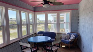 Une véranda avec un ventilateur de plafond, de grandes fenêtres, une table bleue et des meubles en osier.