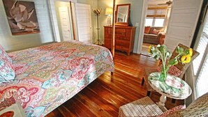 A bedroom with a bed, a dresser, a chair, a table with flowers, and a painting on the wall.