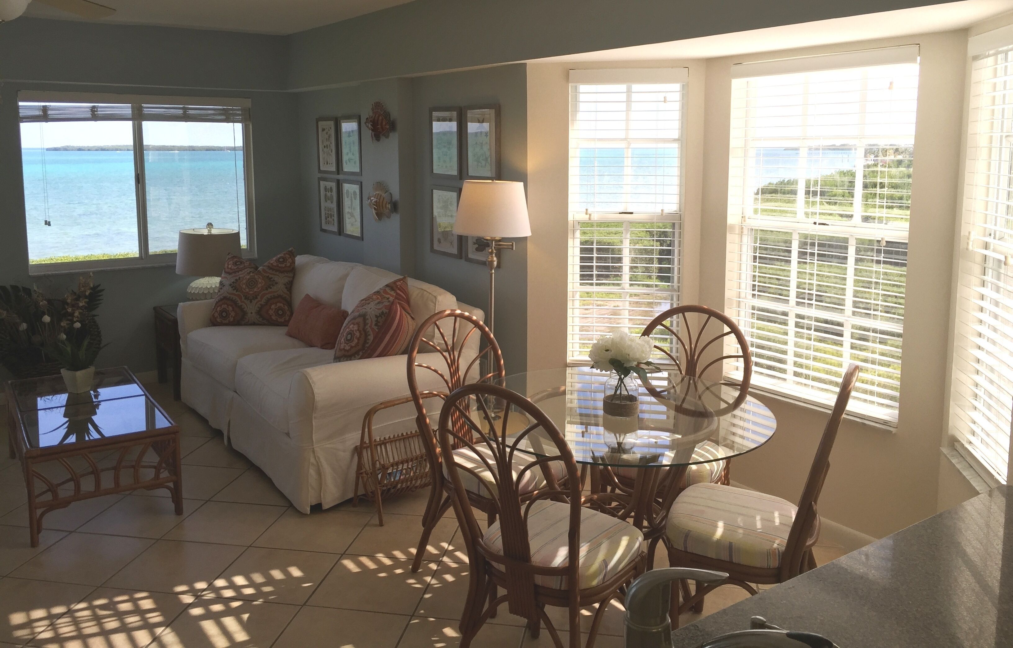 A living room with a view of the sea, a dining area with wicker chairs, and a white sofa with patterned cushions.