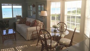 Un salon avec vue sur la mer, un coin repas avec des chaises en osier et un canapé blanc agrémenté de coussins à motifs.