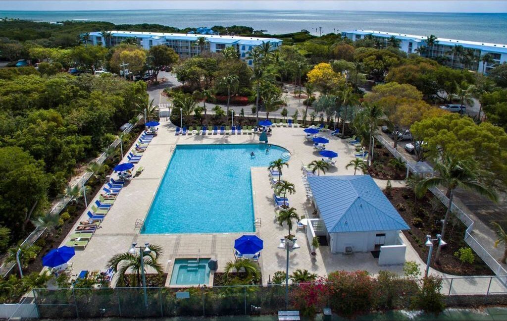 An aerial view of a resort with a central swimming pool, surrounded by lounge chairs and umbrellas, with a backdrop of palm trees and a beach.