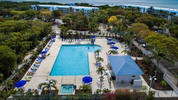 An aerial view of a resort with a central swimming pool, surrounded by lounge chairs and umbrellas, with a backdrop of palm trees and a beach.
