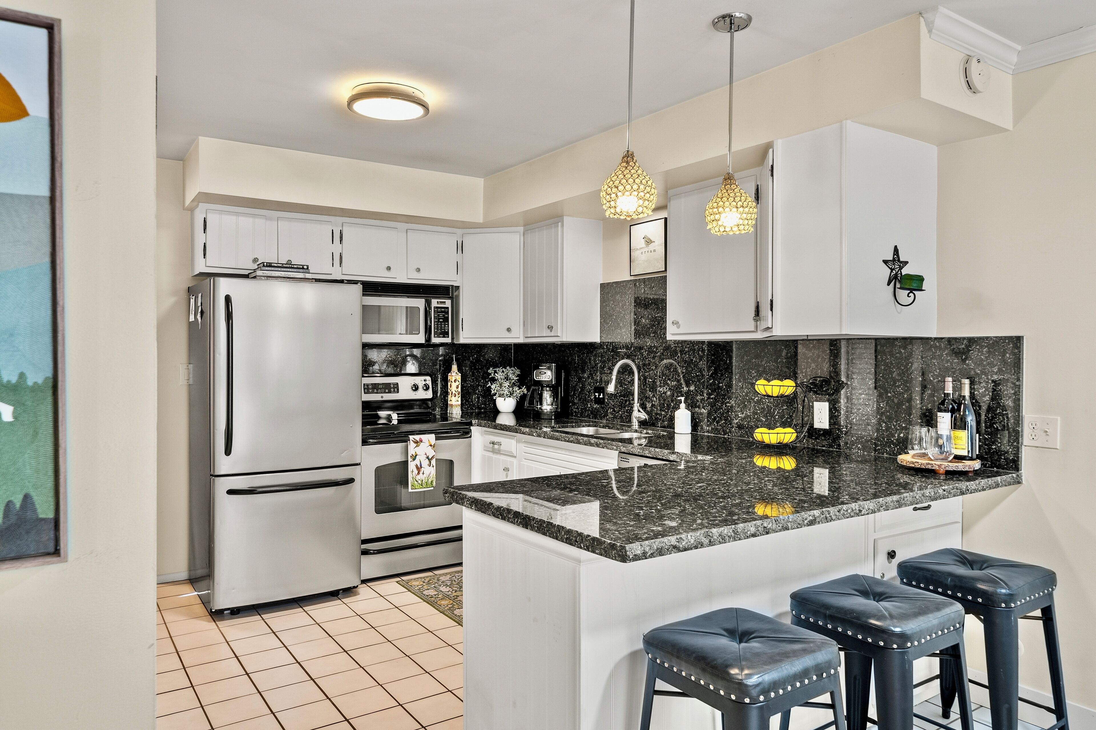 A modern kitchen with white cabinets, a stainless steel refrigerator, a microwave, and a granite countertop. There are three bar stools and pendant lights above the counter.