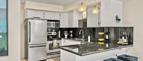 A modern kitchen with white cabinets, a stainless steel refrigerator, a microwave, and a granite countertop. There are three bar stools and pendant lights above the counter.