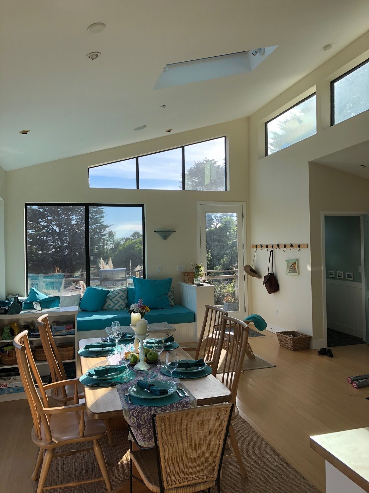 A modern dining area with a high ceiling, large windows offering views of the outdoors, a dining table set with green tableware, and a cozy seating area with a sofa and cushions.