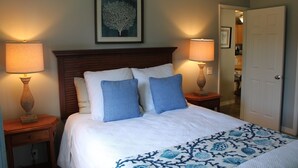 A neatly made bed with a white and blue patterned bedspread, flanked by two bedside tables with lamps, a framed artwork above the bed, and a door to the right. - Plantation Style cottage short walk to beach and town 
Permit STPH2013/0004   (Paia)