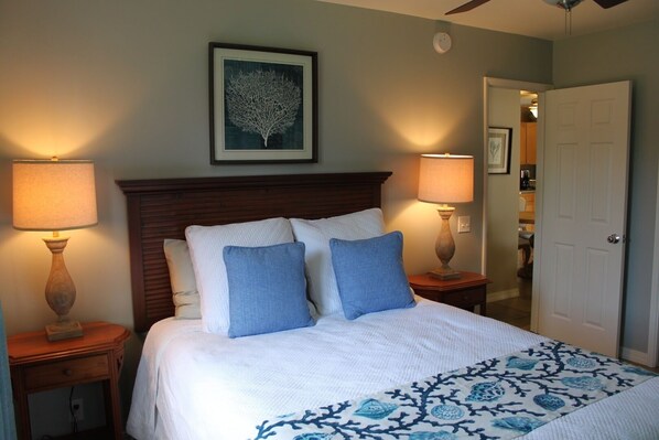 A neatly made bed with a white and blue patterned bedspread, flanked by two bedside tables with lamps, a framed artwork above the bed, and a door to the right. - Plantation Style cottage short walk to beach and town
Permit STPH2013/0004 (Paia)