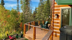 Una terraza de madera con mobiliario exterior, una parrilla y vistas a una zona boscosa.