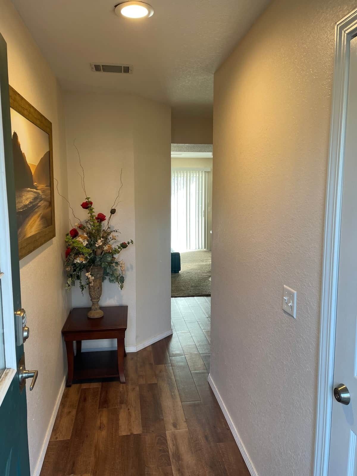 Un pasillo con suelo de madera, un jarrón con flores, un cuadro en la pared y una puerta a la derecha.