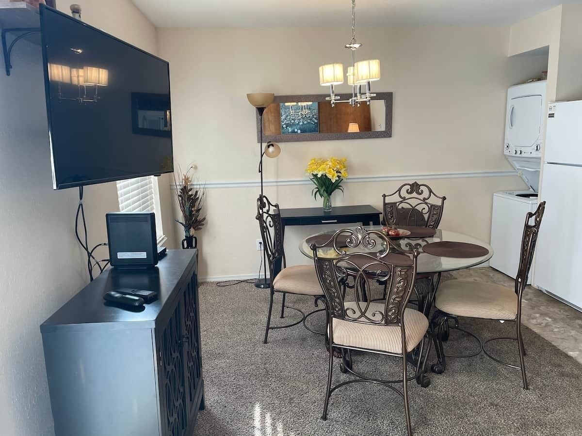 A dining area with a round table, four chairs, a flat-screen TV, a coffee maker, and a refrigerator.