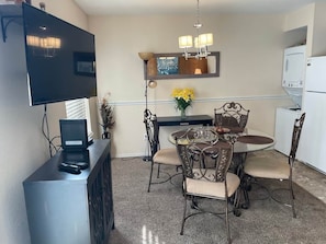 A dining area with a round table, four chairs, a flat-screen TV, a coffee maker, and a refrigerator.