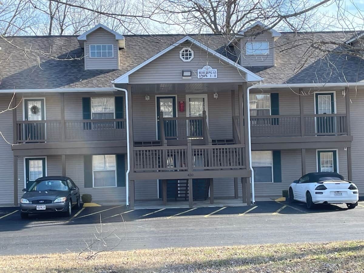 A multi-story building with a central entrance, a balcony, and a parking lot with two cars.