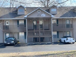 A multi-story building with a central entrance, a balcony, and a parking lot with two cars.