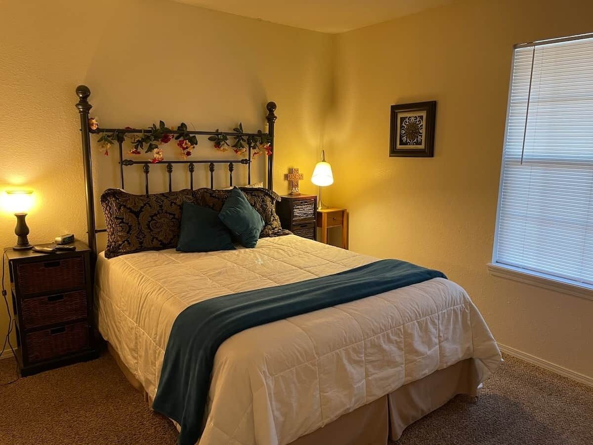 A bedroom with a four-poster bed, two bedside tables, a lamp, and a window with blinds.