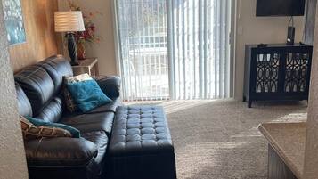 A living room with a leather sofa, a matching ottoman, a TV on a stand, and a large window with blinds.