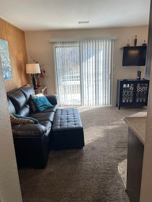 A living room with a leather sofa, a matching ottoman, a TV on a stand, and a large window with blinds.