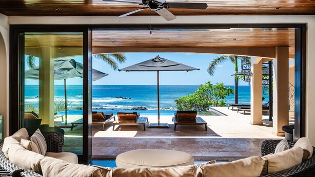A modern lounge area with a view of the ocean, featuring wicker furniture, a ceiling fan, and outdoor umbrellas.