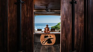 A wooden door leading to a room with a view of the ocean, a meditation statue, and a unique wooden table.