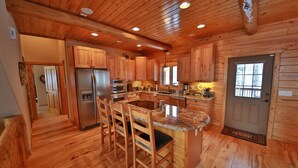 A wooden cabin interior with a kitchen featuring a refrigerator, oven, and wooden cabinets. There are three wooden chairs around a granite countertop island.
