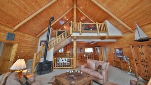 A spacious log cabin interior with a high ceiling, a central staircase, a cozy living area with a sofa and coffee table, and a fireplace.