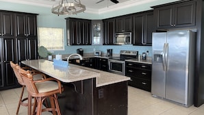 A kitchen with dark cabinets, a stainless steel refrigerator, a microwave, a stove, and a ceiling fan. - The Beach House - Panoramic Ocean Views & Beach Access Near Local Attractions (Orange Beach)