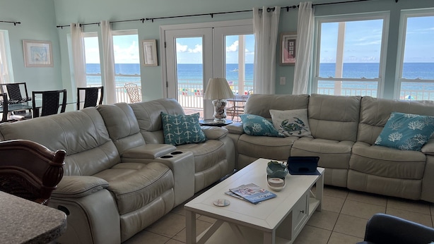 A spacious living room with a large beige sectional sofa, a white coffee table, and a view of the ocean through large windows. - The Beach House - Panoramic Ocean Views & Beach Access Near Local Attractions (Orange Beach)