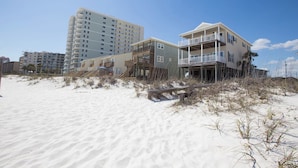 A beachfront property with a wooden deck and a balcony, surrounded by sand and sparse vegetation. - The Beach House - Panoramic Ocean Views & Beach Access Near Local Attractions (Orange Beach)