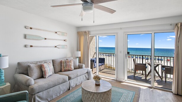 Un salon avec un canapé, une table basse et un ventilateur de plafond. Il y a un balcon avec vue sur la plage et l’océan.