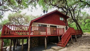 A red wooden cabin with a stone foundation, a wooden deck, and a staircase leading to a balcony. - Casa Rio Hideaway @ Casa Rio Rentals- Pet Friendly with River access (Concan)