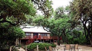 A stone building with a red exterior and a balcony, surrounded by trees and a stone fire pit with chairs. - Casa Rio Hideaway @ Casa Rio Rentals- Pet Friendly with River access (Concan)