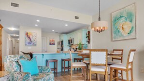 Un salon spacieux avec un canapé à motifs, des chaises en bois et un coin repas, sur fond d’une cuisine bleu clair.
