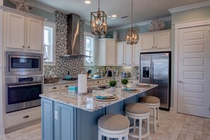 Een moderne keuken met witte kasten, een centraal eiland met krukken, roestvrijstalen apparatuur en decoratieve schelpen op de bovenkasten.