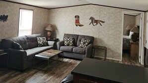 En stue med en grå sofa, en mindre grå lænestol og et sofabord. Der er en lampe på et sofabord, og en loftsvifte. Væggene er beige med en struktureret overflade.