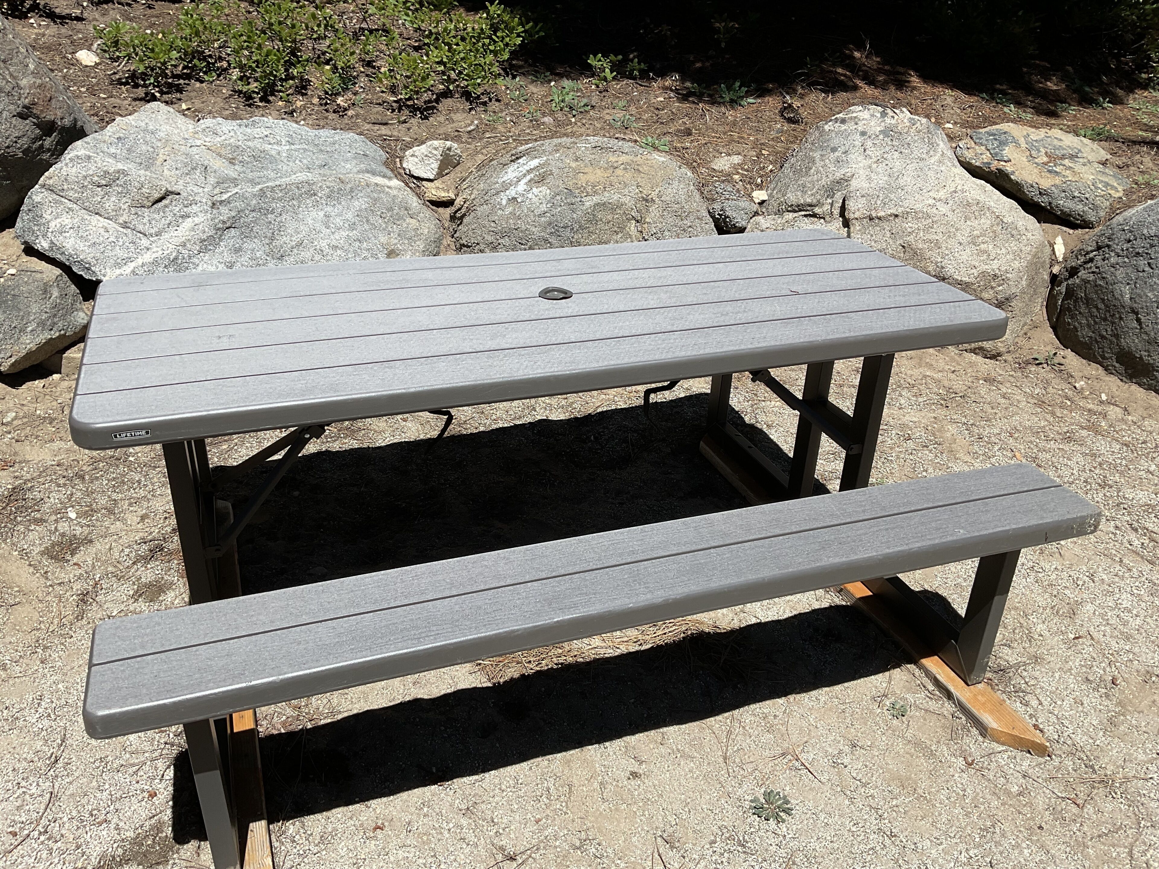 Une table de pique-nique en métal gris, avec un banc, installée à l’extérieur sur une surface de gravier, avec de gros rochers en arrière-plan.